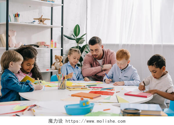 教师和种族学龄前儿童在桌上与油漆和纸在教室里
